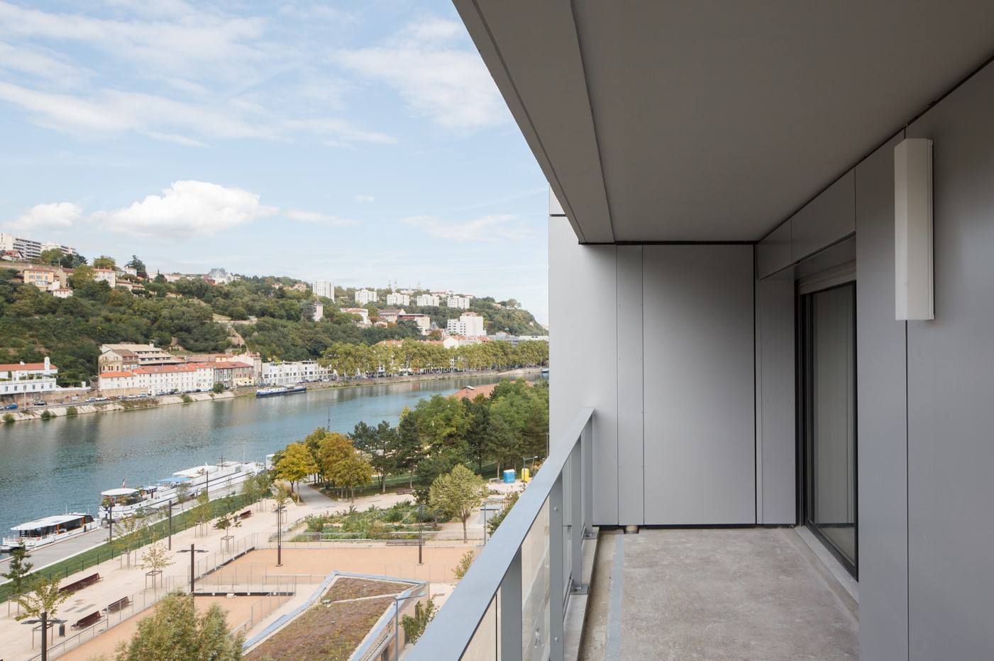 Vue depuis un balcon moderne sur les quais de Saône à Lyon, avec des bâtiments colorés en bord de rivière et une colline boisée à l’arrière-plan.