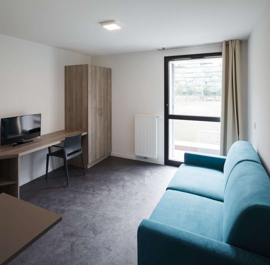 Studio étudiant lumineux avec un canapé turquoise, un bureau équipé, une armoire en bois, une fenêtre et une porte vitrée donnant sur l’extérieur.