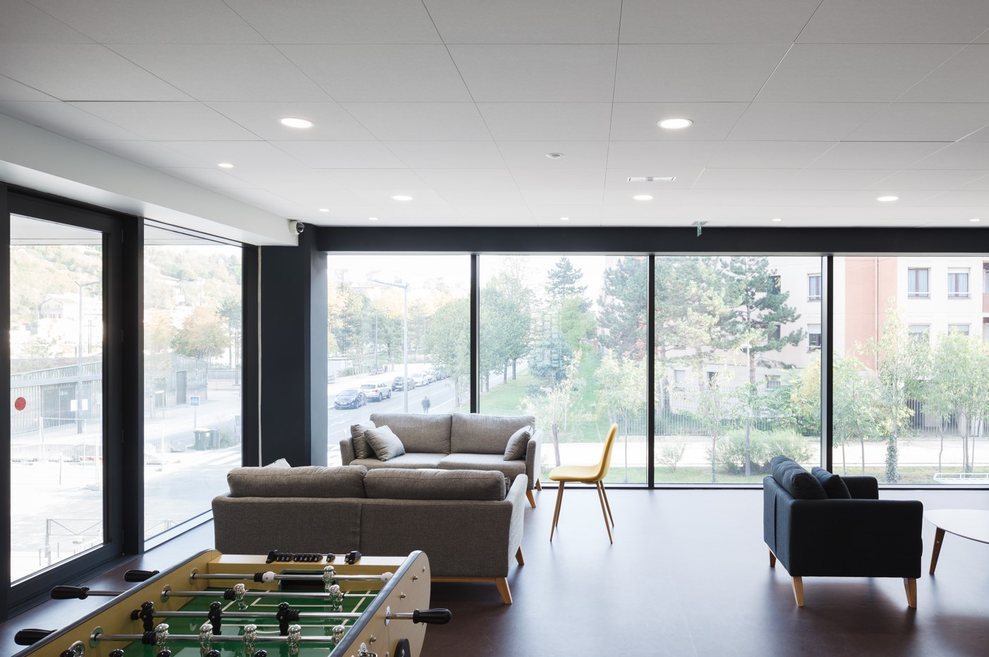 Espace commun lumineux avec de larges baies vitrées, canapés gris, fauteuils, une chaise jaune et un babyfoot en premier plan.
