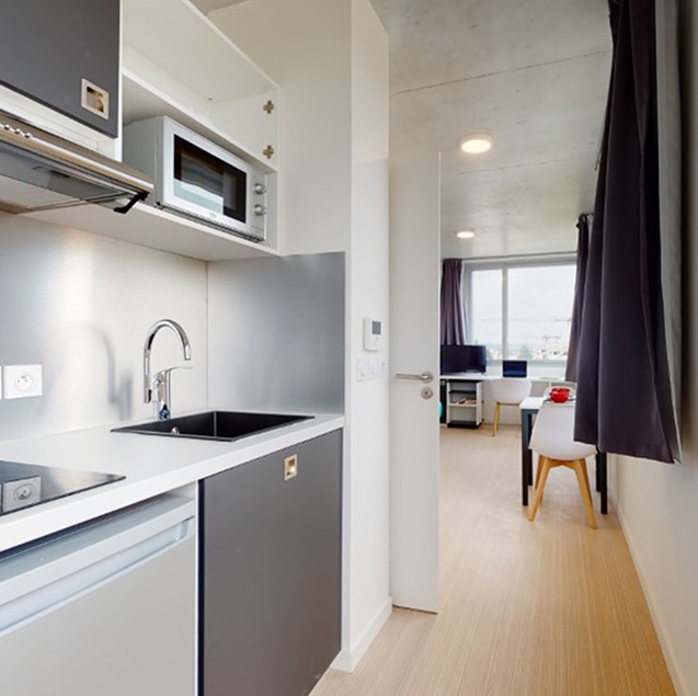 Intérieur d’un studio étudiant avec kitchenette équipée, plaques de cuisson, évier, micro-ondes, et vue sur un coin bureau lumineux à l’arrière-plan.