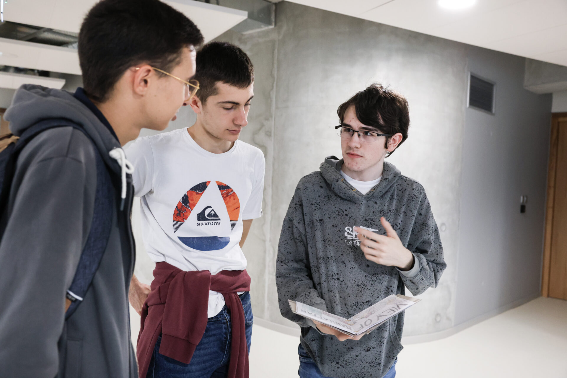 Trois étudiants discutent dans un couloir en tenant un carnet de croquis ou un projet artistique, en contexte scolaire.