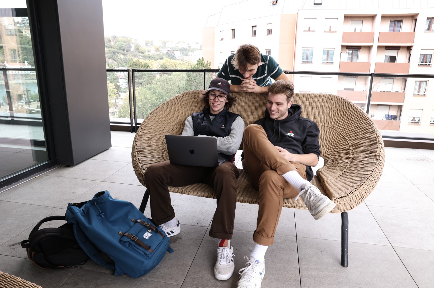 Trois étudiants assis sur une banquette en rotin sur une terrasse, regardent ensemble un ordinateur portable, dans une ambiance détendue.