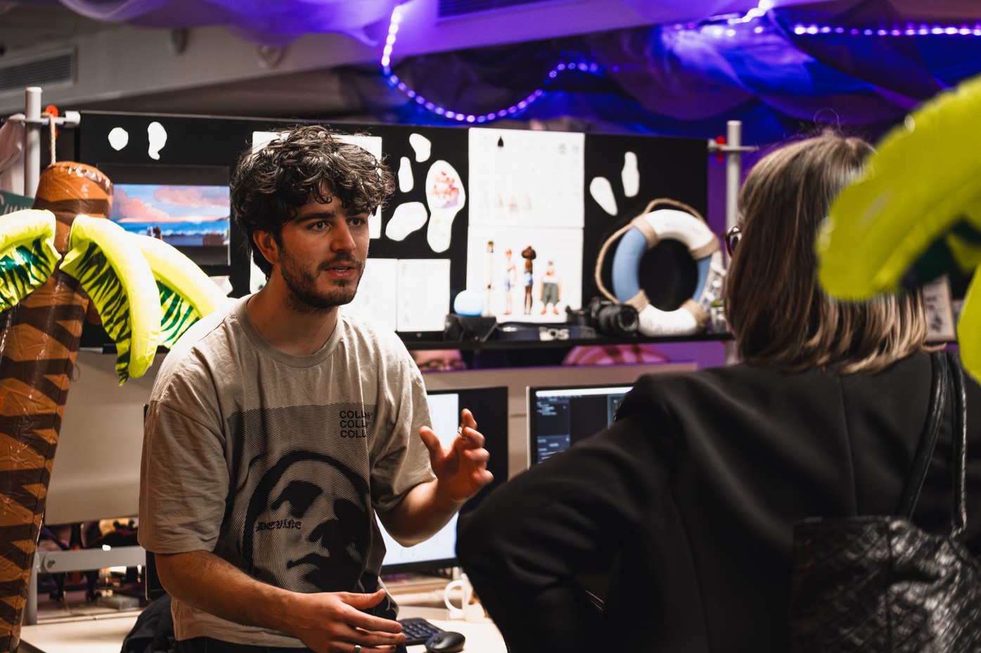 Un étudiant en cinéma d’animation 3D présente son projet à une visiteuse, dans un espace décoré de palmiers gonflables et de planches de recherche visuelle.
