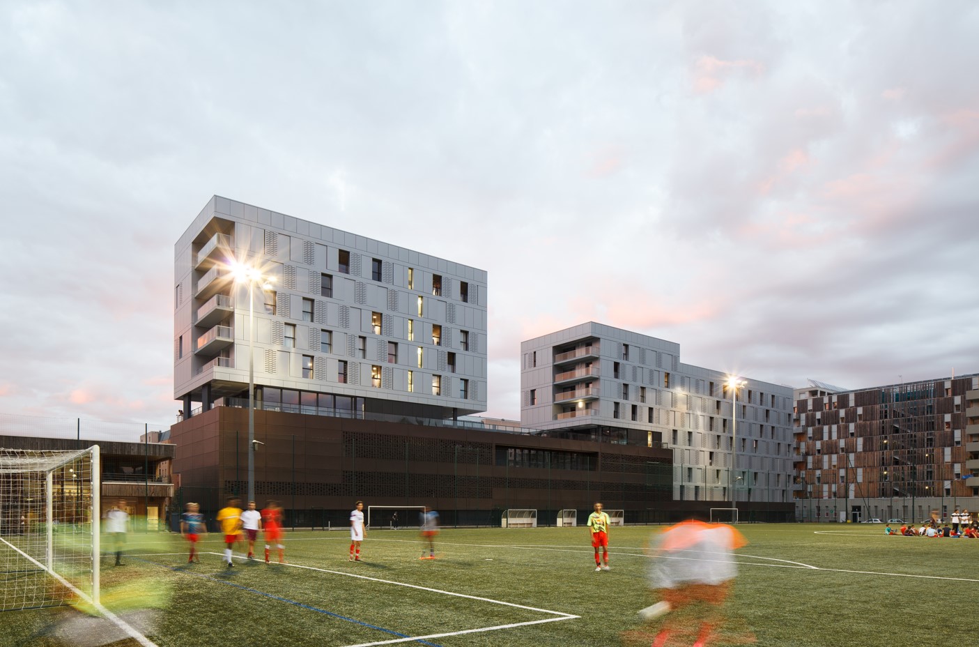 Vue d’un terrain de football avec des étudiants en mouvement, en soirée, devant un ensemble de bâtiments modernes et éclairés.