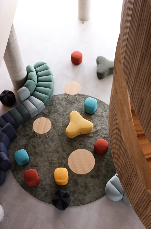 Vue plongeante sur un hall lumineux avec mobilier design coloré, canapés modulaires, poufs et tables rondes sur un grand tapis circulaire.
