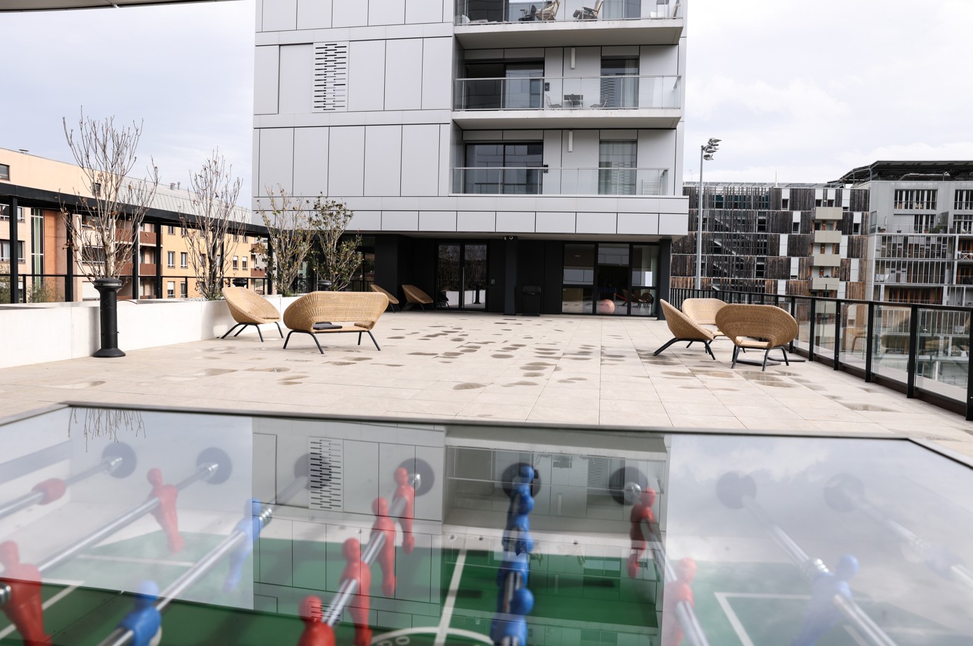 Grande terrasse extérieure aménagée avec fauteuils en rotin, vue sur des bâtiments modernes, avec un babyfoot en premier plan.