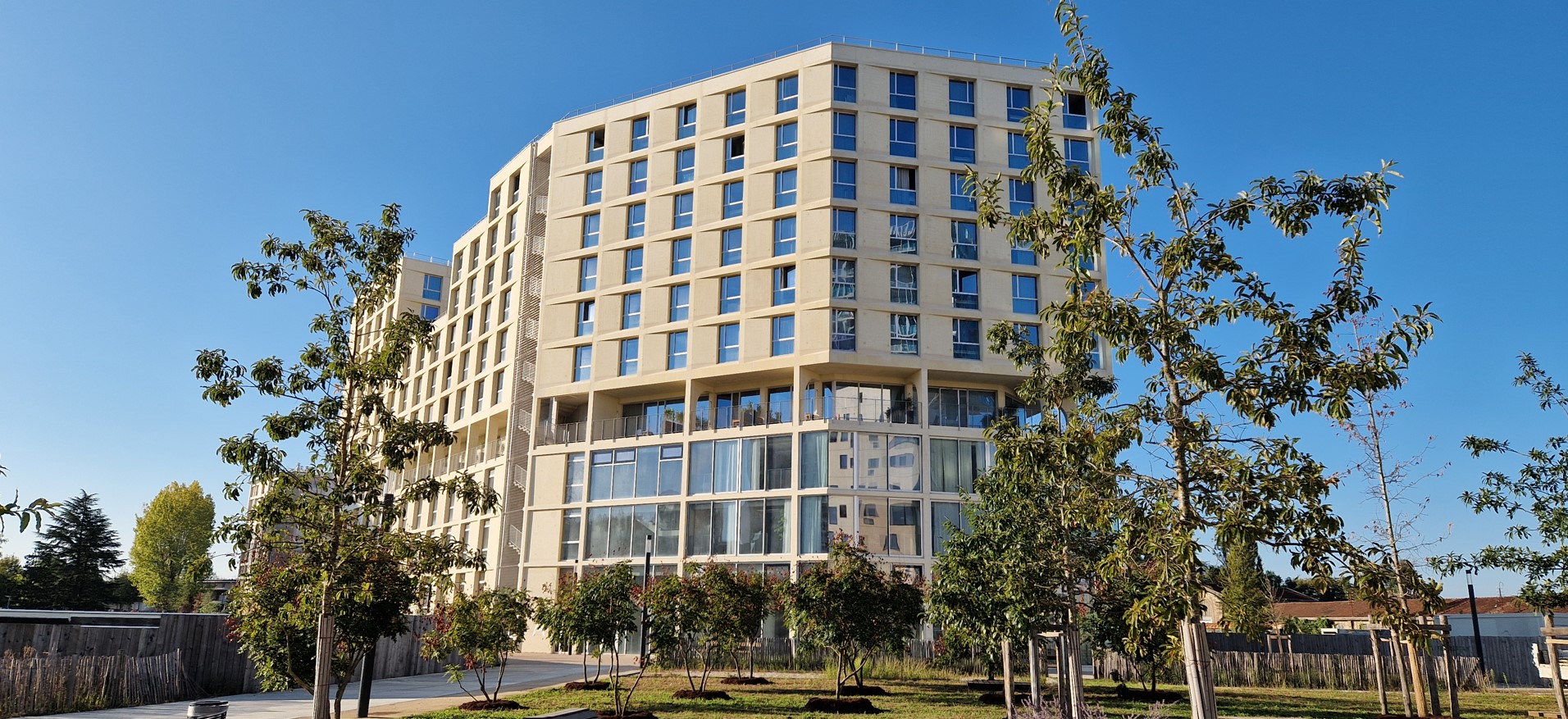 Grand bâtiment moderne à plusieurs étages avec de nombreuses fenêtres, entouré d’un jardin arboré sous un ciel bleu.